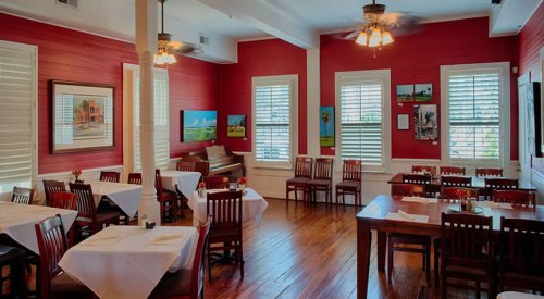 Cozy restaurant interior with wooden tables, red walls, and artwork, featuring bright windows and a warm atmosphere.