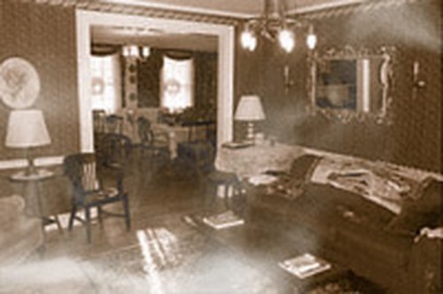 A vintage living room with a sofa, lamps, and a mirror, featuring patterned wallpaper and a dining area in the background.