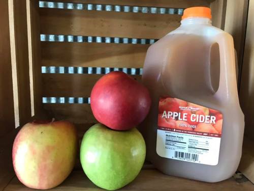 A jug of apple cider next to three apples: one red, one green, and one yellow, arranged on a wooden crate.