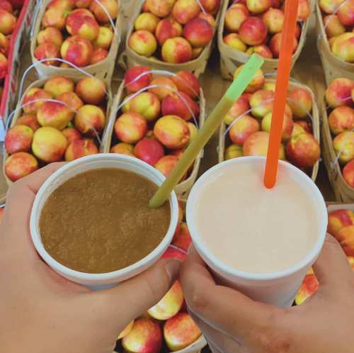 Two hands holding cups with drinks, one brown and one white, in front of baskets filled with red apples.