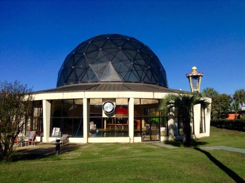 A geodesic dome building with large windows, surrounded by green grass and palm trees, under a clear blue sky.