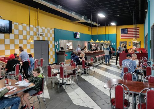 A colorful diner interior with booths, tables, and patrons enjoying meals, featuring an American flag on the wall.