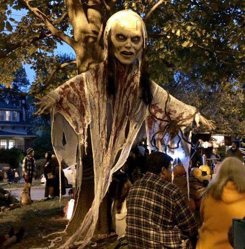 A spooky ghost figure with long hair and tattered clothing hangs from a tree, surrounded by people in a Halloween setting.