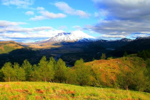 A scenic view of snow-capped mountains under a blue sky, surrounded by green hills and valleys.
