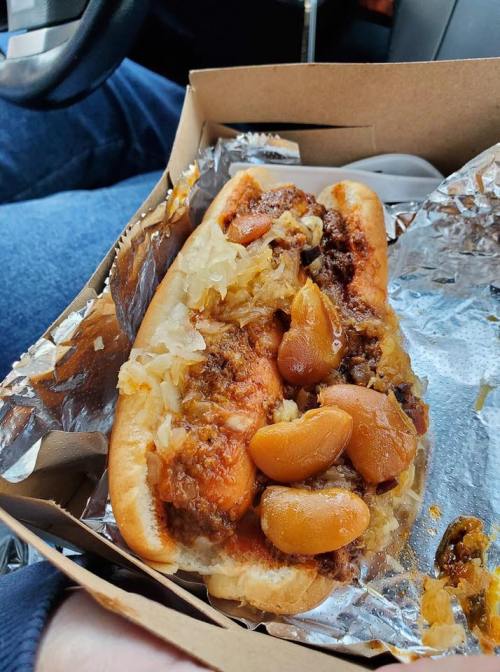 A close-up of a hot dog topped with chili, onions, and mustard, served in a takeout box.