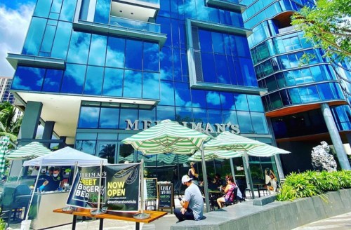 Modern building with large glass windows, outdoor seating, and striped umbrellas. Sign reads "Now Open."
