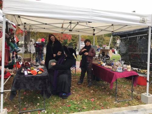 Three vendors at a market booth under a white tent, showcasing various crafts and decorations. Fall foliage in the background.