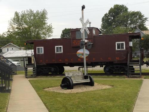 A vintage caboose on display near a railroad crossing sign, surrounded by grass and a bench in a park setting.