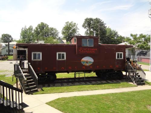 A vintage red caboose with "Victoria Station" and "Sugar Creek RR" signage, set on a grassy area with steps leading up.