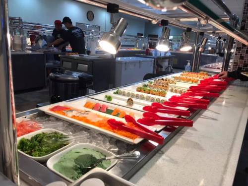A sushi buffet with various sushi rolls, sashimi, and green seaweed salad, served with red tongs in a well-lit setting.