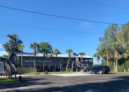 A rustic building with palm trees, located in a sunny area, featuring a parking lot and stairs leading to the entrance.