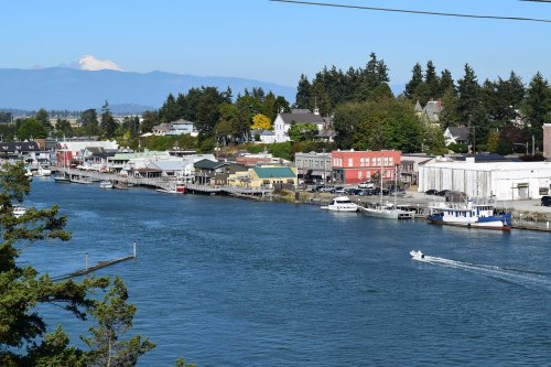 A scenic view of a waterfront with boats, shops, and trees, set against a backdrop of distant mountains.