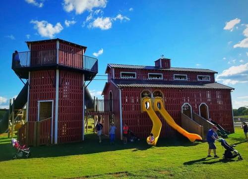 A colorful playground resembling a barn, featuring slides and a tower, with children and adults enjoying the space.