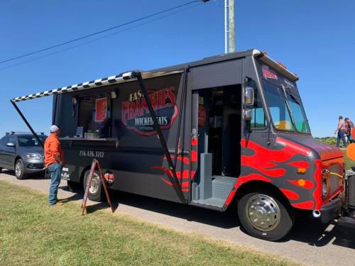 A food truck with a black and red flame design, parked on a sunny day, serving hot dogs and snacks.