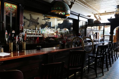 A dimly lit bar with a polished wooden counter, shelves of liquor, and decorative antlers on the wall.