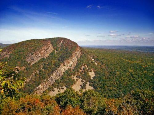 A panoramic view of a lush, green mountain landscape under a clear blue sky with scattered clouds.