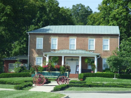 A two-story brick house with a green metal roof, surrounded by lush greenery and colorful flower pots on a wagon.