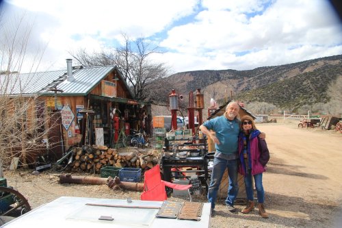 A man and woman stand together outside a rustic building surrounded by vintage equipment and scenic mountains.