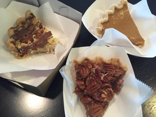 Three slices of pie on white paper: chocolate pecan, pumpkin, and traditional pecan, displayed on a black table.