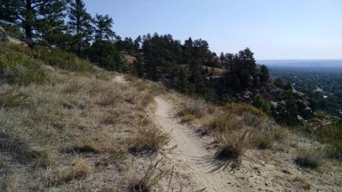 A winding dirt path through dry grass and shrubs, surrounded by trees and a distant view of a valley.