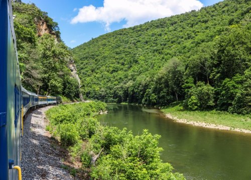 A scenic view of a train traveling along a river, surrounded by lush green hills and blue skies.