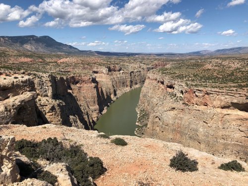 A scenic view of a canyon with a winding river, surrounded by rocky cliffs and a blue sky with scattered clouds.