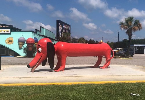 A large, red dog sculpture with floppy ears stands on a concrete platform under a blue sky.
