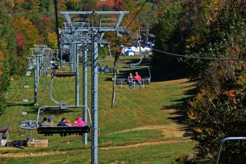 A scenic view of a ski lift with people riding, surrounded by colorful autumn foliage and a grassy area below.