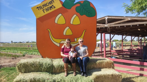 Two women sit on hay bales in front of a large pumpkin decoration at Tolmachoff Farms, holding a baby.