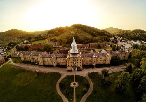 Aerial view of a historic building with a clock tower, surrounded by green hills and a small town at sunset.
