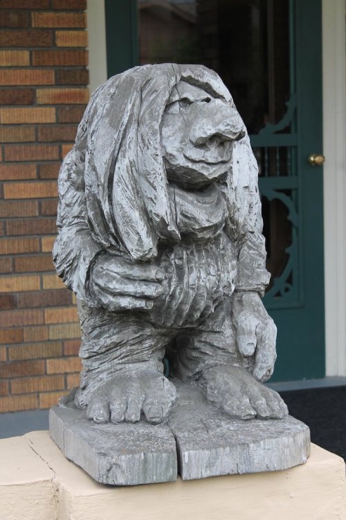 A weathered stone statue of a troll-like creature with long hair, standing on a pedestal in front of a brick building.