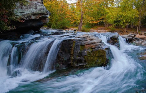 A serene waterfall cascades over rocks, surrounded by vibrant autumn foliage and a tranquil forest setting.