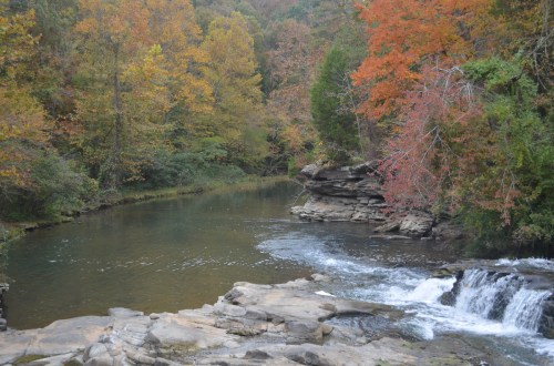 A serene river flows through a forest with vibrant autumn foliage in shades of orange and red.