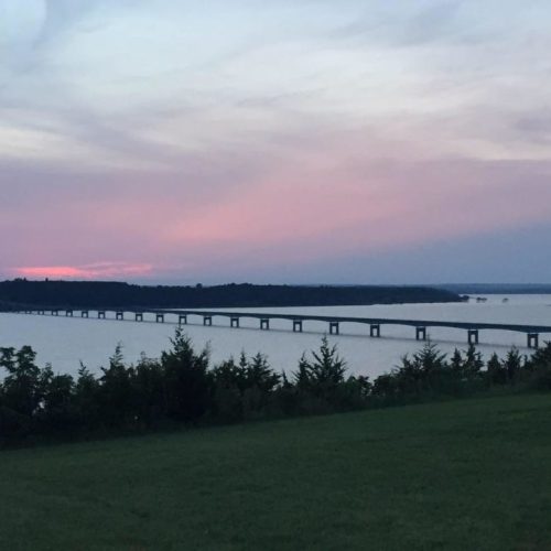 A serene sunset over a bridge spanning a calm body of water, with lush greenery in the foreground.