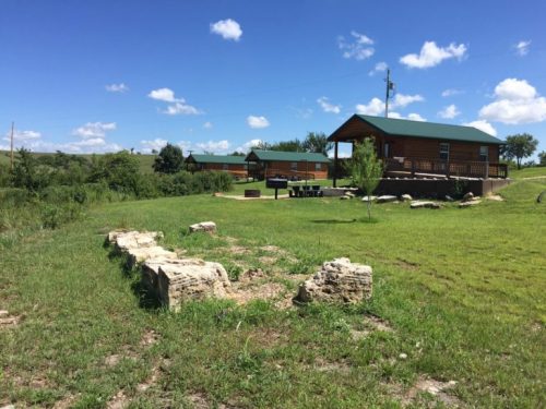 A grassy area with stone remnants and two wooden cabins under a blue sky with scattered clouds.