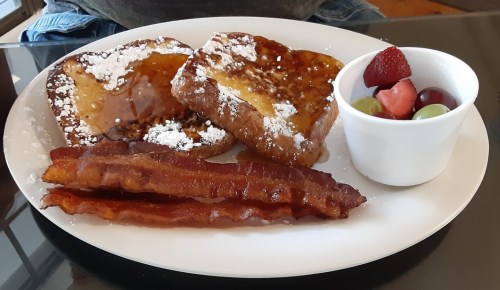 A plate with French toast dusted with powdered sugar, crispy bacon, and a small cup of mixed fruit.