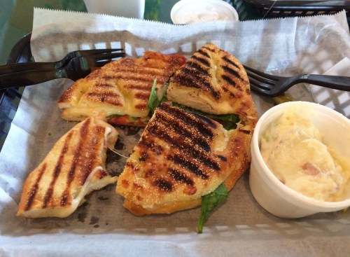 Grilled sandwich with chicken, spinach, and cheese, served with a side of creamy potato salad in a takeout basket.