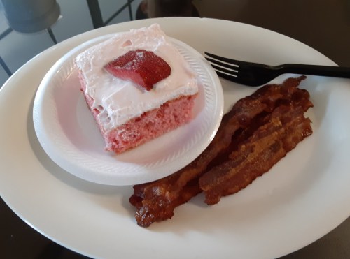 A slice of strawberry cake with a strawberry on top, served with crispy bacon on a white plate.