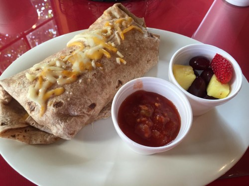 A plate with a cheese-topped burrito, salsa, and a cup of mixed fruit including grapes, pineapple, and a strawberry.