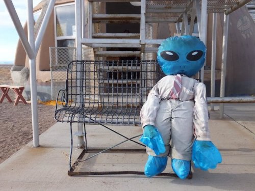 A blue alien figure in a suit sits on a metal bench outside a building in a desert landscape.