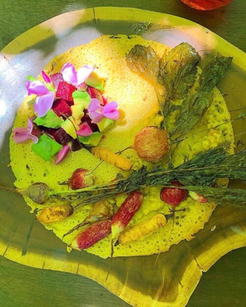 A vibrant dish featuring a yellow pancake topped with fresh fruits, edible flowers, and herbs on a wooden plate.