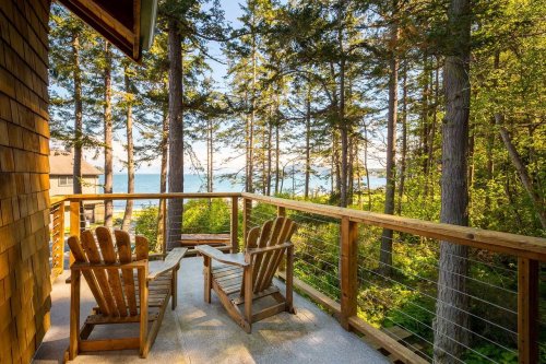 A serene balcony with two wooden chairs overlooking a forest and a tranquil body of water in the distance.