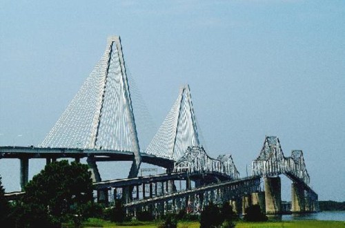 A modern bridge with distinctive triangular towers spans a river under a clear blue sky.