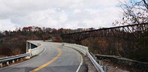 A winding road curves alongside a bridge, surrounded by autumn trees and a cloudy sky.