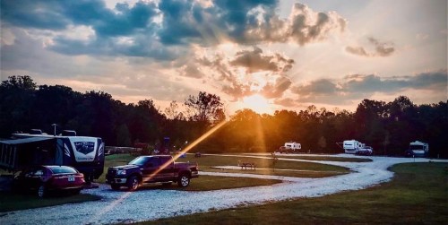 A serene campsite at sunset, with RVs parked along a winding gravel path and trees in the background.