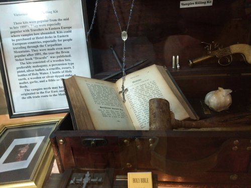 A display case containing a vampire killing kit with a book, cross, wooden stake, and a silver bullet.