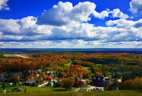 A scenic view of colorful autumn trees, a valley, and a blue sky with clouds, overlooking a small town and ski lifts.