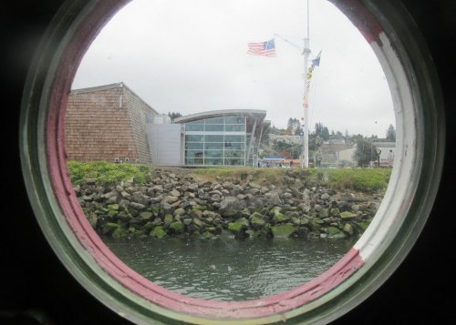 View through a circular window of a building by the water, with rocks and a flag in the background.