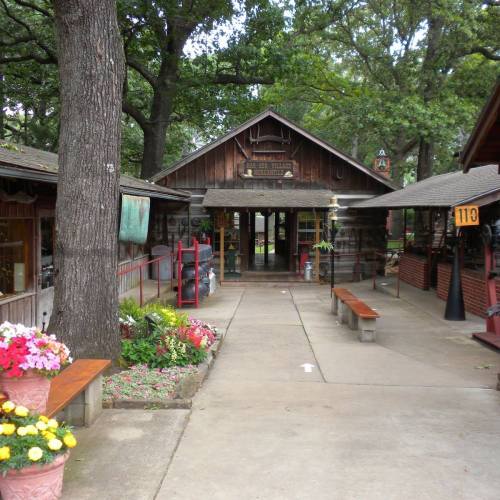 A rustic wooden building with a sign, surrounded by flower pots and benches, set in a tree-lined area.
