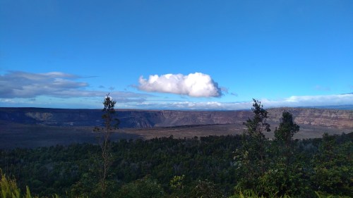 A panoramic view of a volcanic landscape with a blue sky and a single cloud above, framed by green trees.